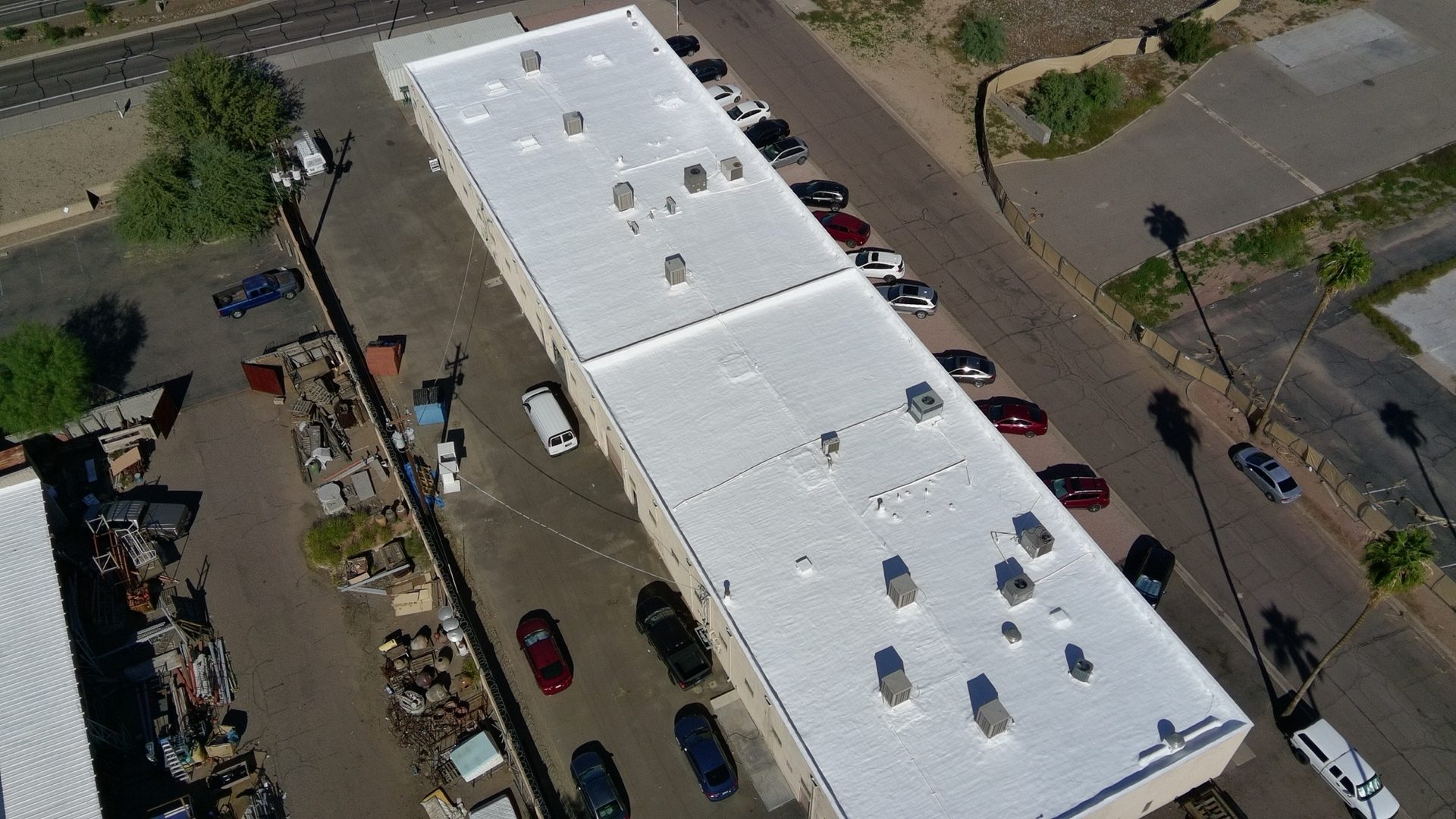 Aerial drone view of the completed 16,600 sq ft new spray polyurethane foam roof at 116 S Industrial Dr in Tempe AZ finished bright white with two coats of ARMORCOAT AC100 elastomeric coating, palm shadows across the street and the industrial corridor of west Tempe visible below