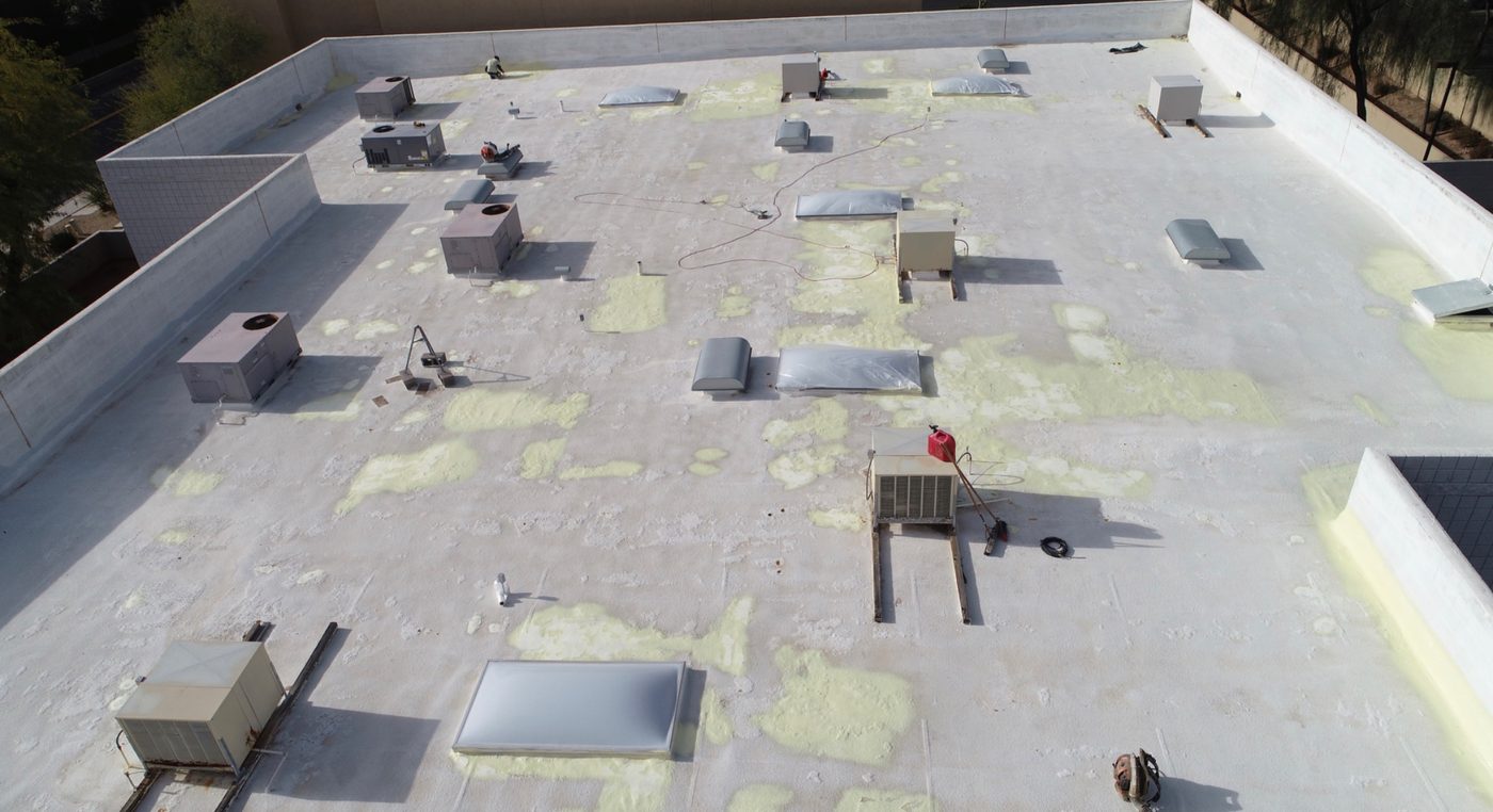 During the job: fresh yellow spray polyurethane foam patches over primed gray substrate on the Chandler industrial roof, showing blister cut-out and refill work and low-area buildup with a worker on the roof