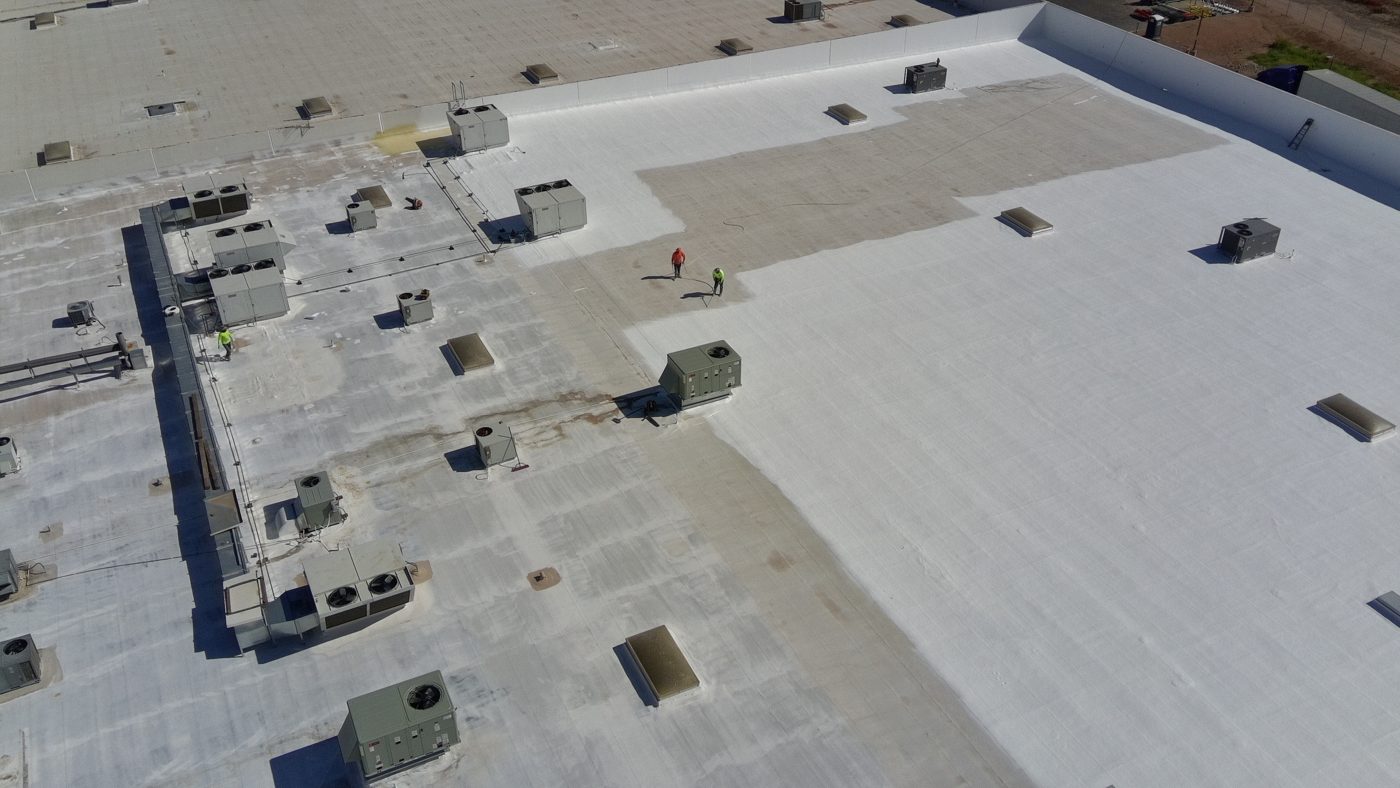 During the job: aerial drone view of fresh bright white ARMORCOAT AC100 coating being applied on the Tempe warehouse foam roof next to the old gray weathered surface, with crew on the roof