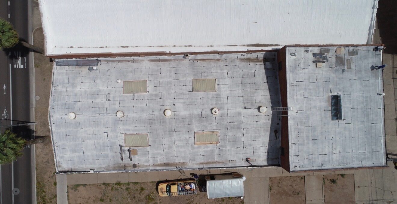 Before restoration: top-down aerial view of the PBF Manufacturing Phoenix warehouse showing an aged silver-coated cap-sheet modified bitumen roof with 4 old yellowed fiberglass skylights and multiple weathered vent penetrations