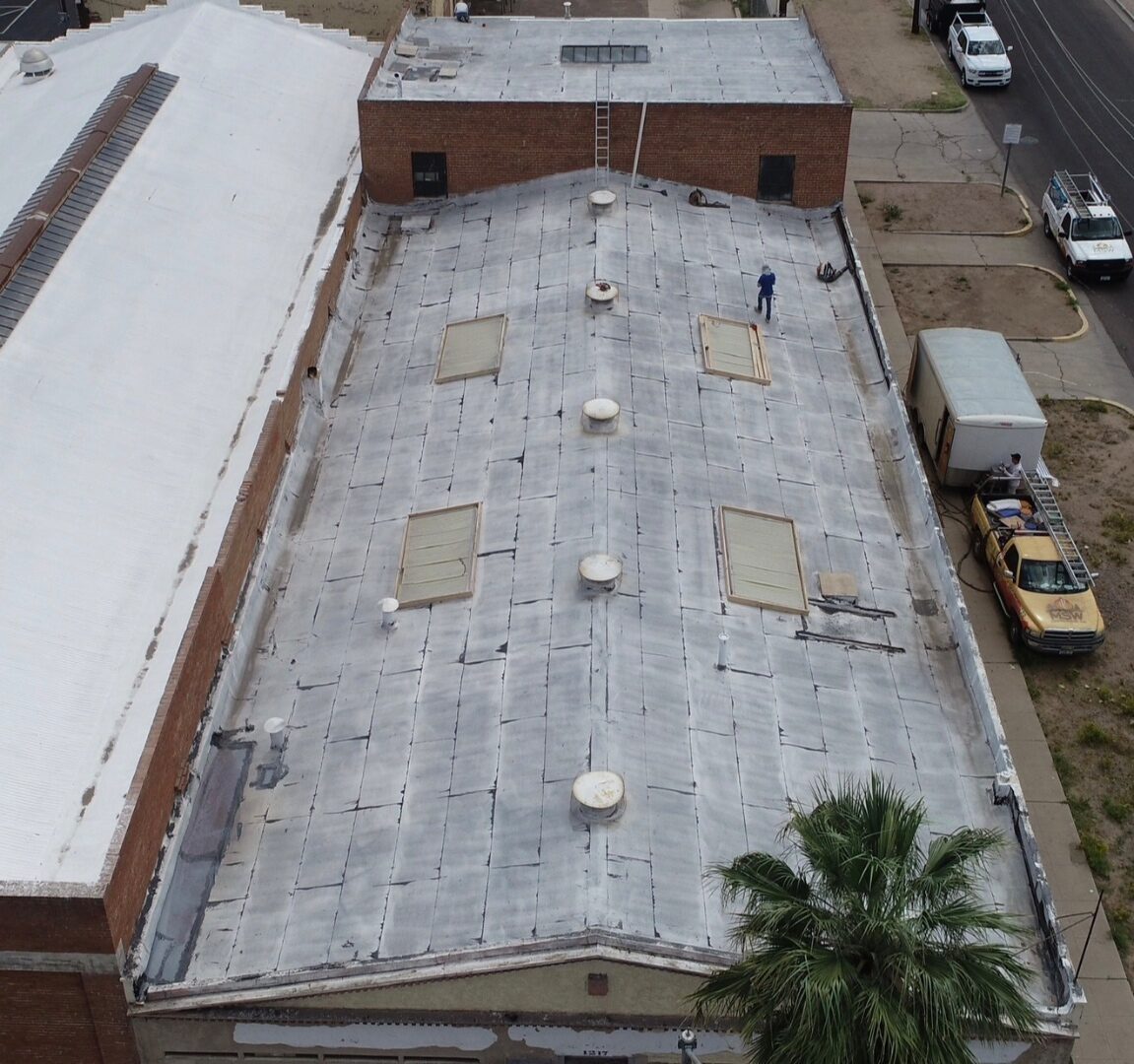 Before restoration: shallow drone angle of the aged silver-coated cap-sheet roof at PBF Manufacturing Phoenix with an MSW Vanguard Roofing box truck staged on the street below the warehouse