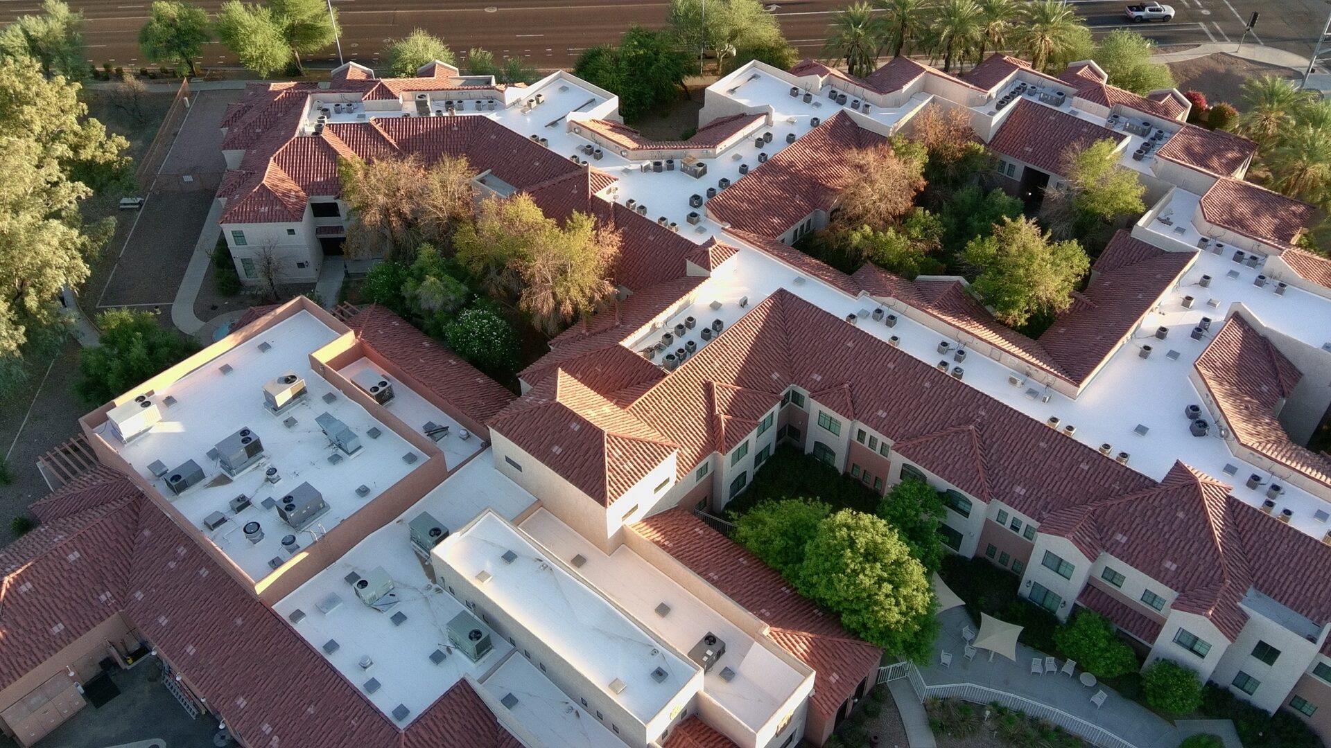 Aerial drone view of the completed 20,000 sq ft silicone-coated low-slope roof at Solterra Senior Living, 350 S Alma School Rd in Chandler AZ, showing bright white TRC 980 100% silicone coating running the length of the assisted-living community between the terracotta Spanish-tile pitched wings, HVAC units cleanly detailed across the field, and the Alma School corridor visible in the background
